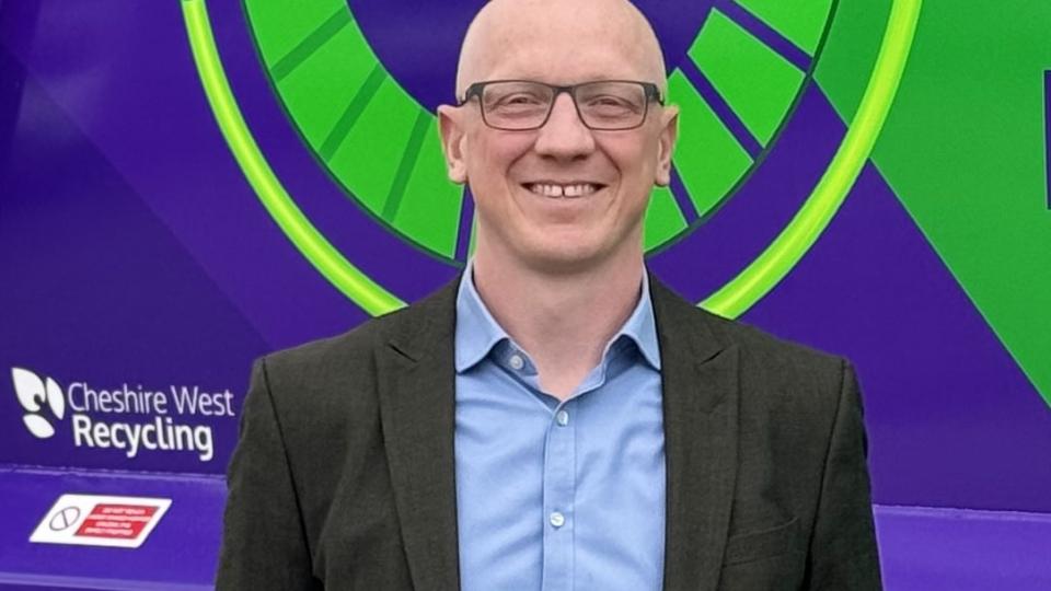 A man with glasses, blue shirt and black suit jacket stands in front of a purple refuse truck with green icon.